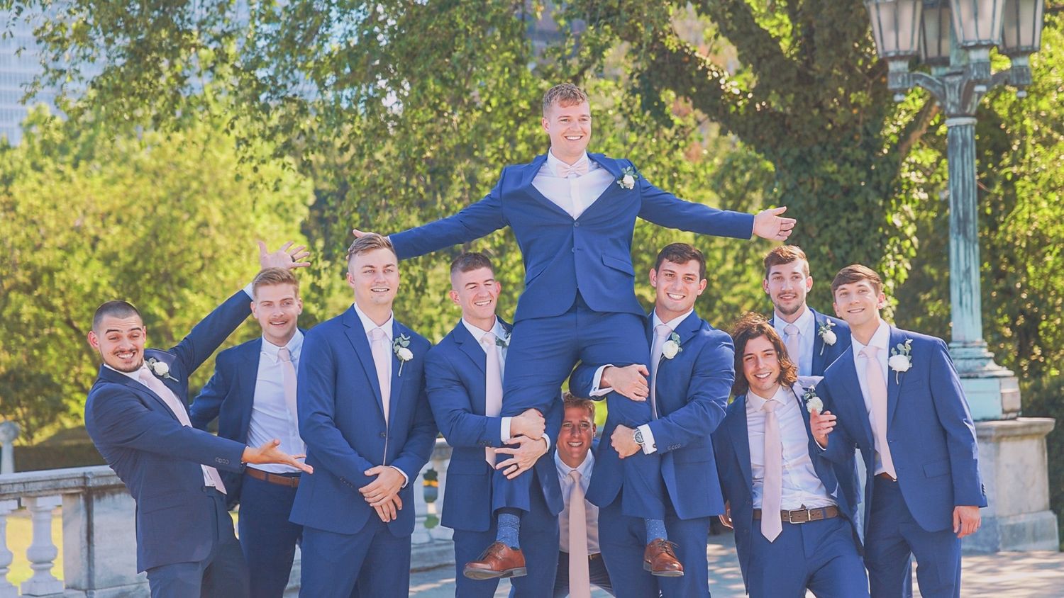 A cheerful group in coordinated navy suits lifts one person wearing a bright blue sweater during an outdoor photo session.