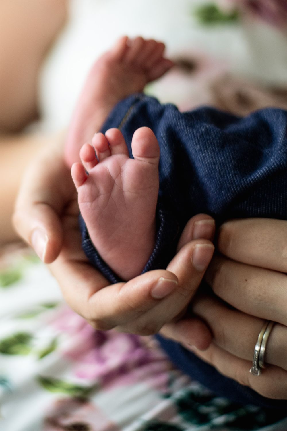 Tiny newborn baby feet being gently cradled in adult hands wearing a wedding ring against floral background.