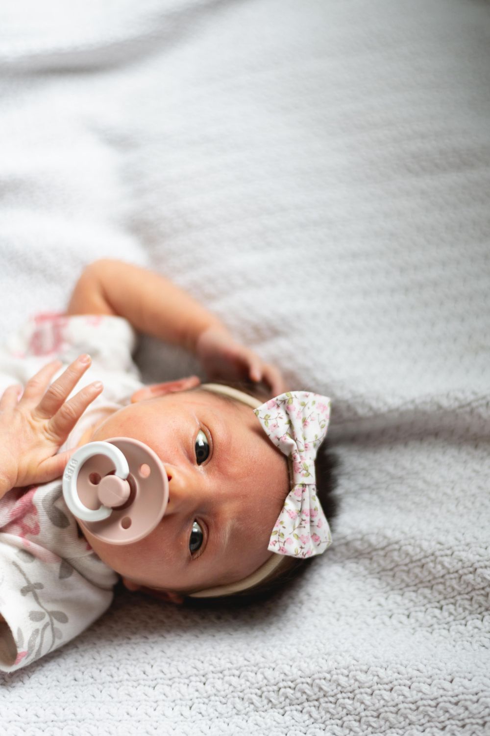 Baby wearing white outfit with floral pattern and bow headband lies on gray blanket with pacifier.