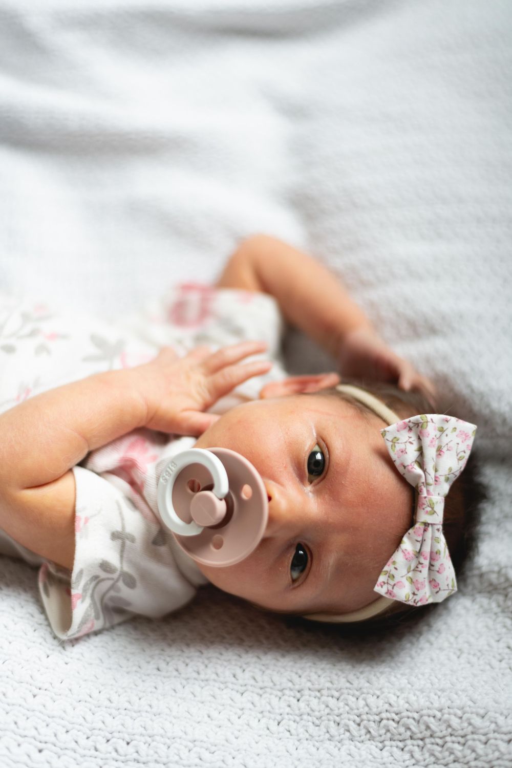 Baby wearing white outfit with floral bow headband laying on grey blanket.