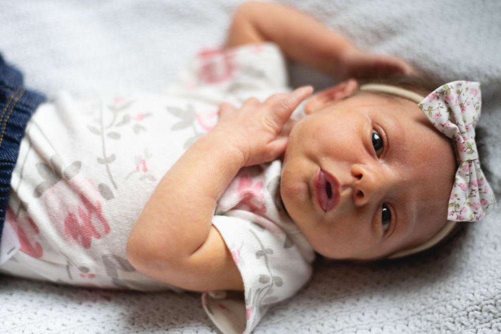 Baby wearing floral outfit and bow lying on grey textured blanket.