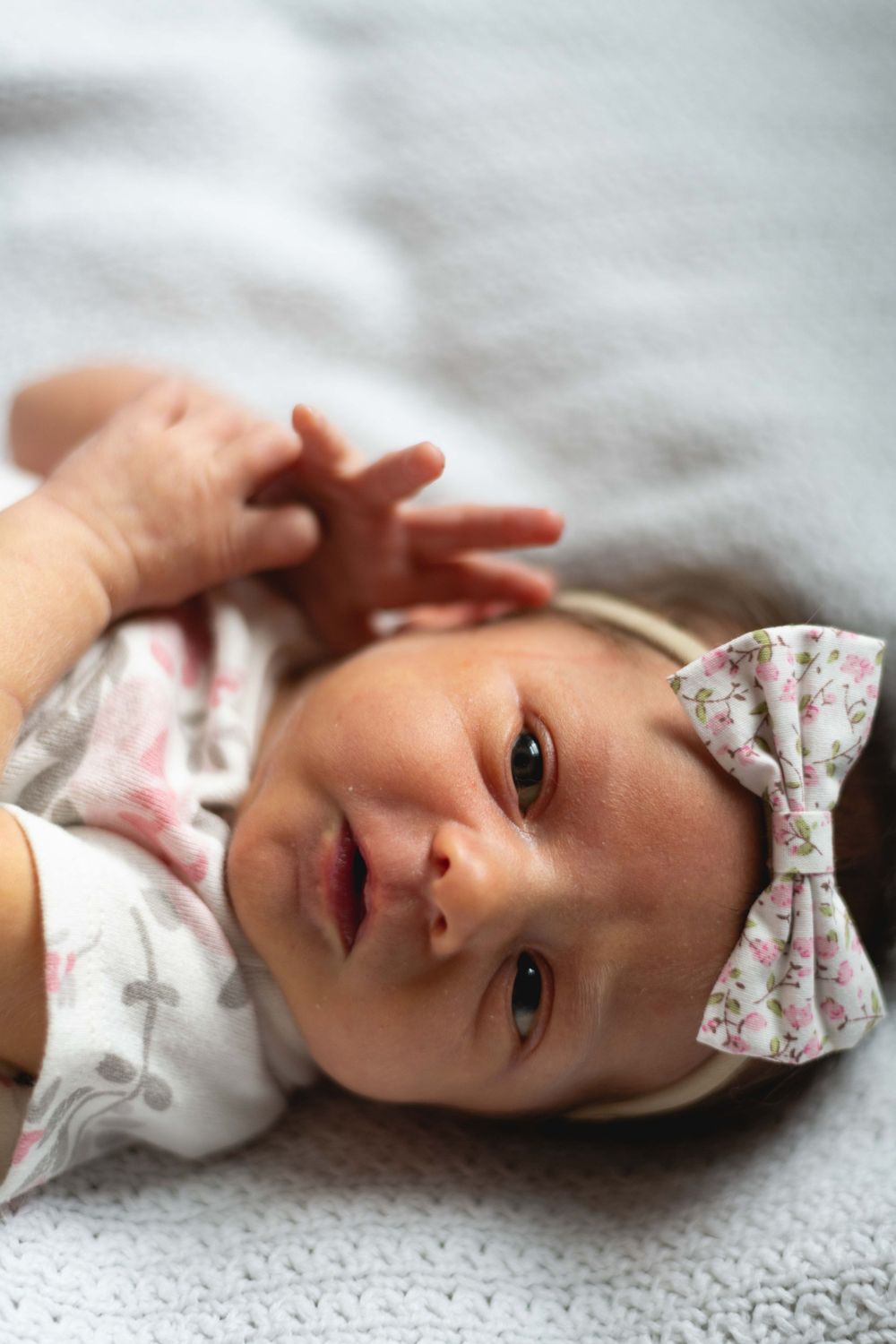 Close up of infant wearing floral dress and bow headband on grey blanket.