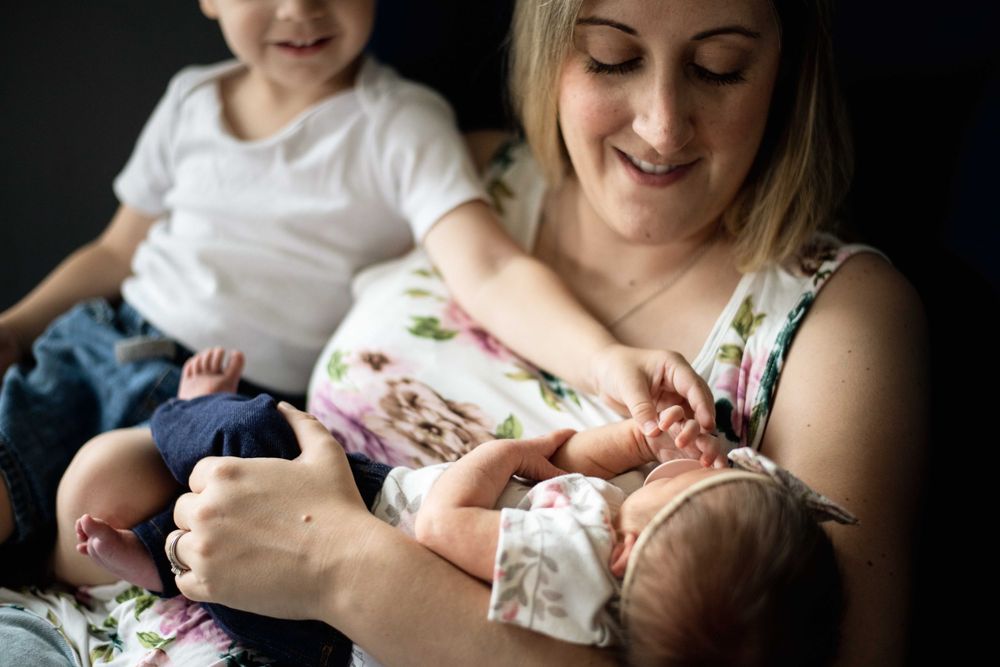 Mother in floral dress tenderly holding newborn while toddler sits nearby.