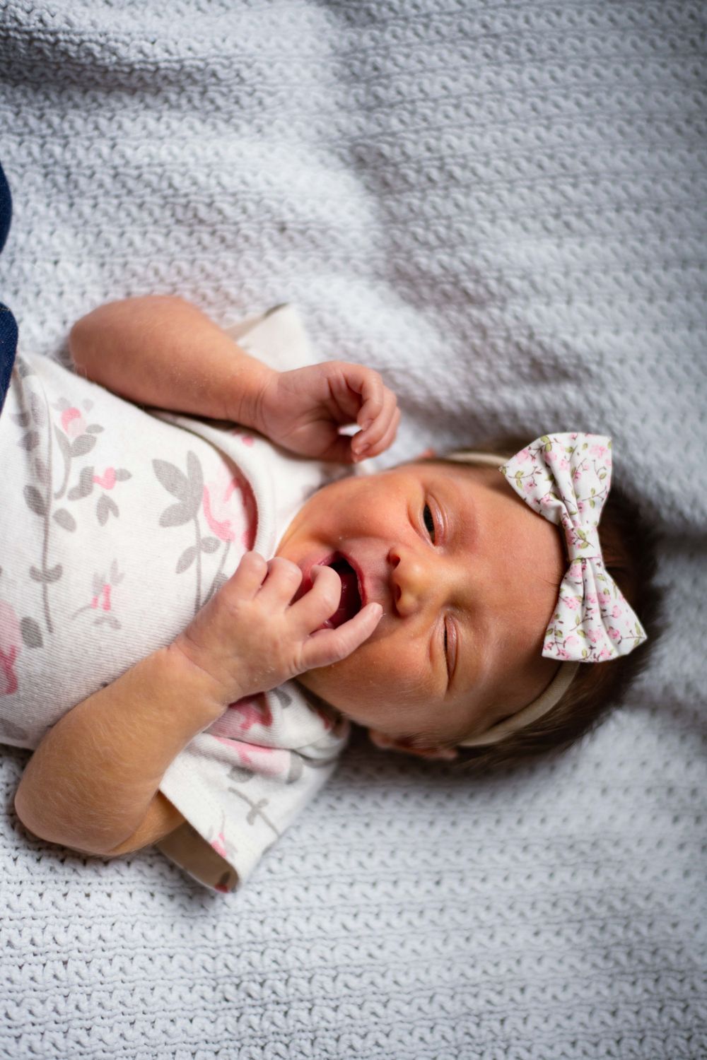 Baby with bow headband lying on textured grey blanket making playful expressions.