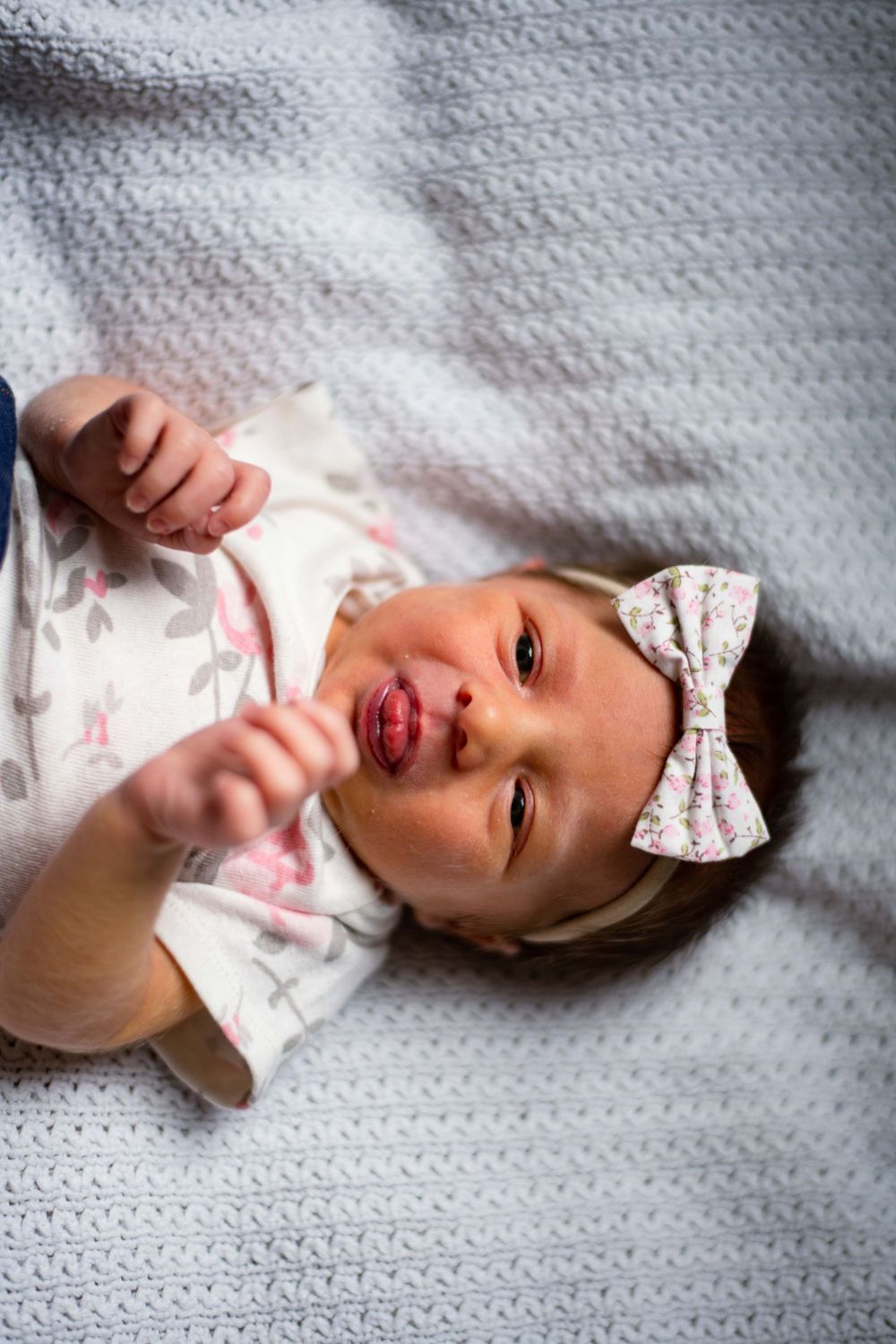 Smiling baby wearing white floral outfit and bow headband on grey blanket.