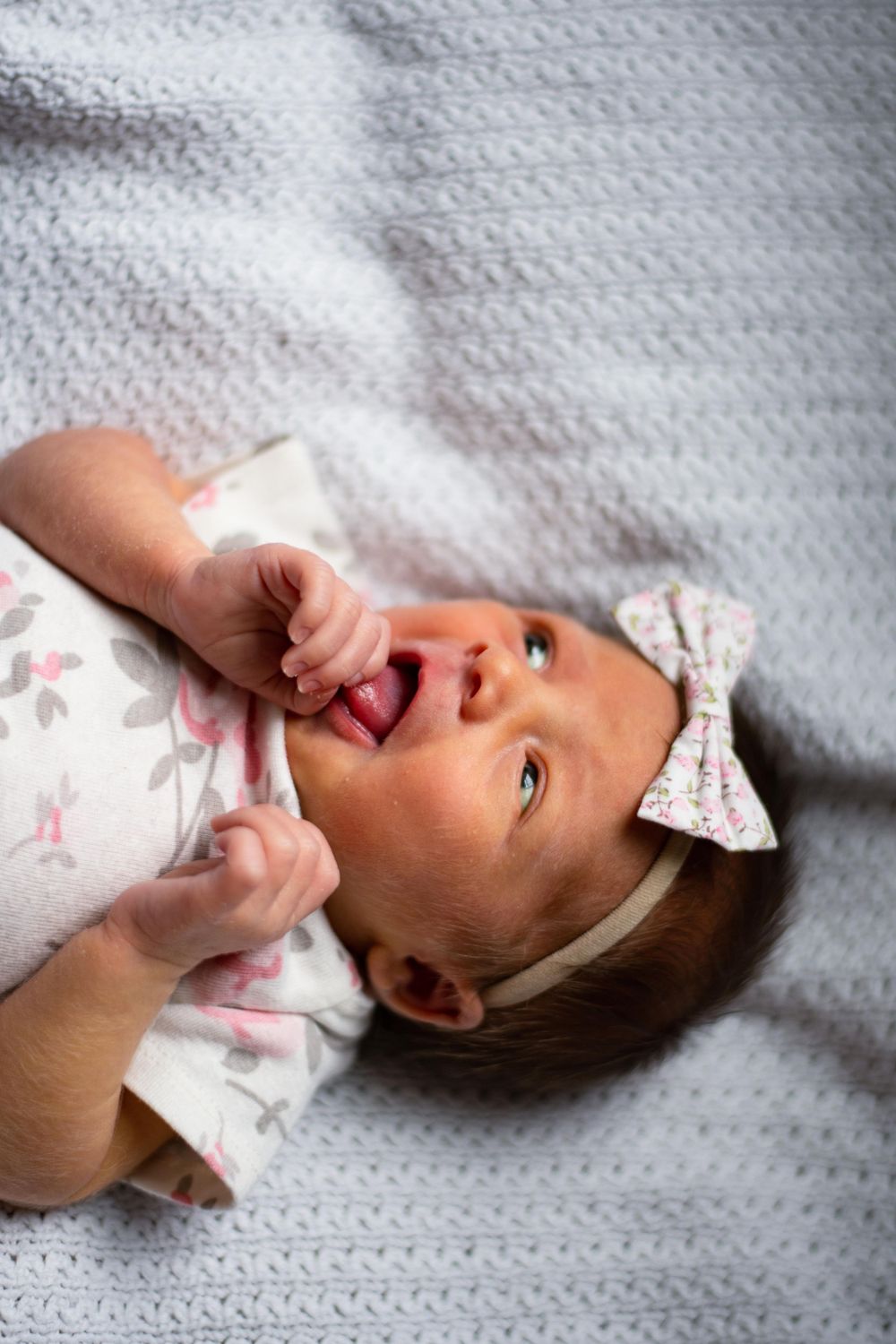 Laughing baby with bow accessory playing while lying on grey textured fabric.