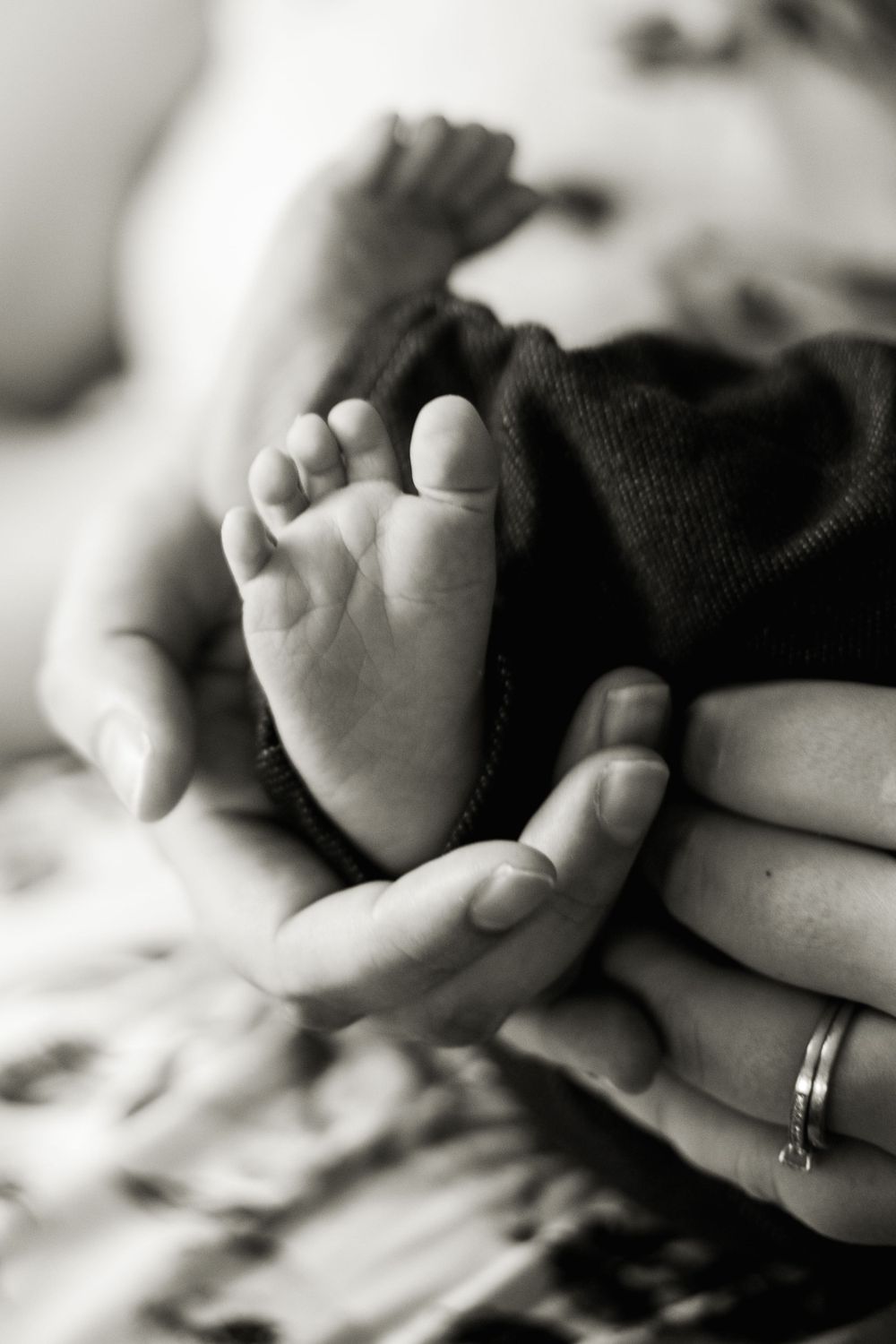 Black and white close-up of tiny newborn foot cradled in adult hands with wedding ring visible.