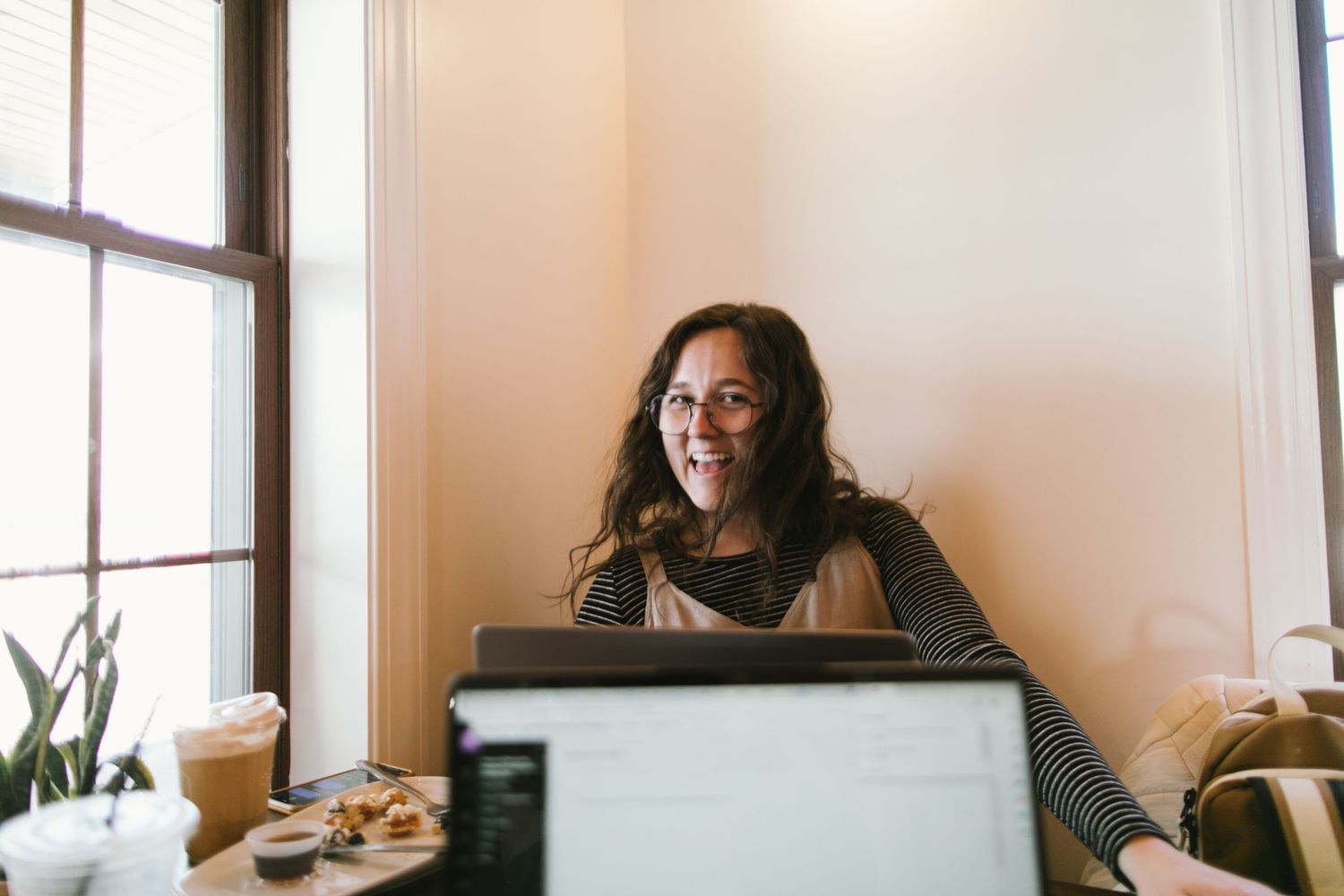 Person working on laptop at table with coffee and snacks near window.