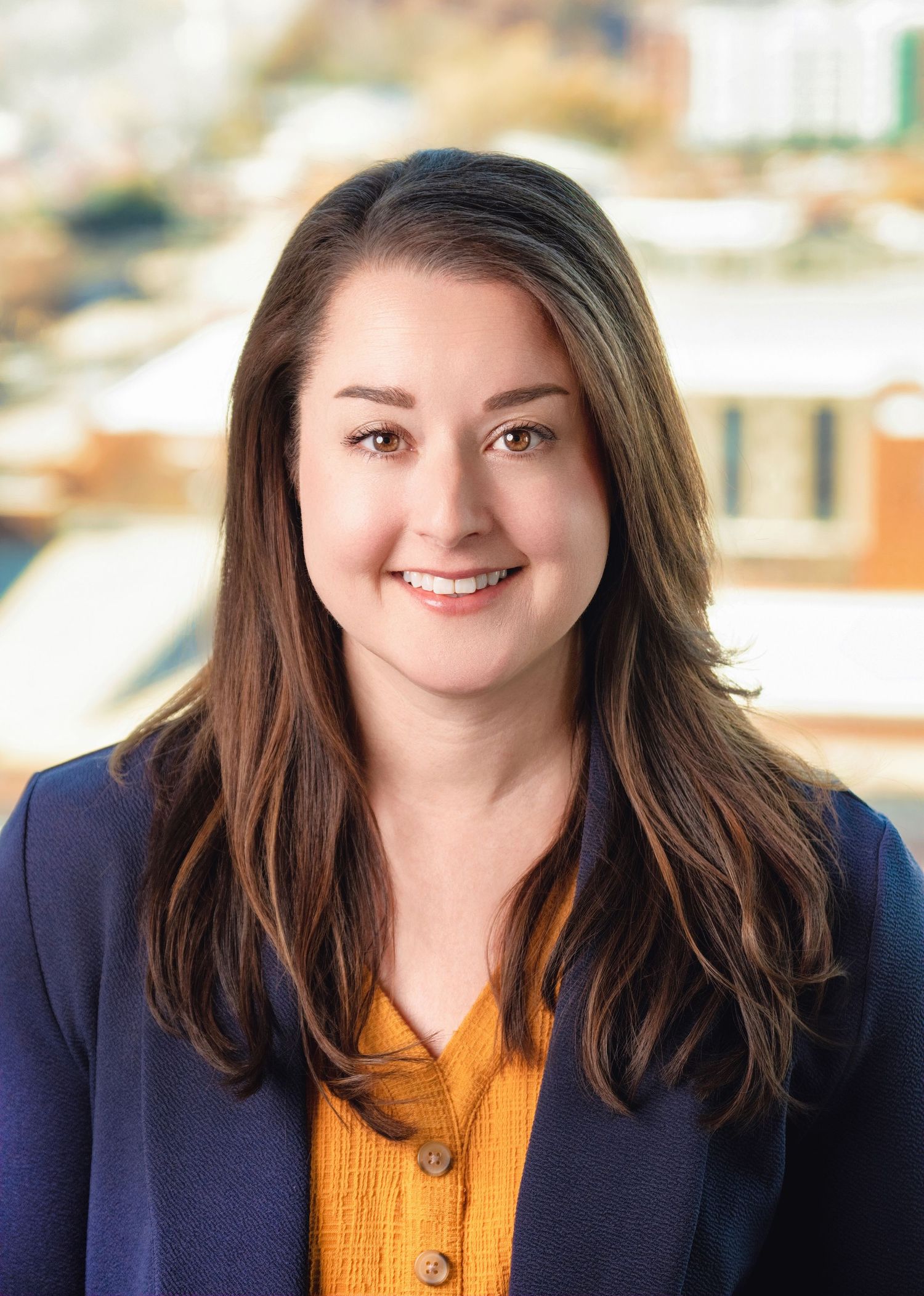 Professional headshot of a person wearing a navy blazer and yellow blouse against a blurred office background.