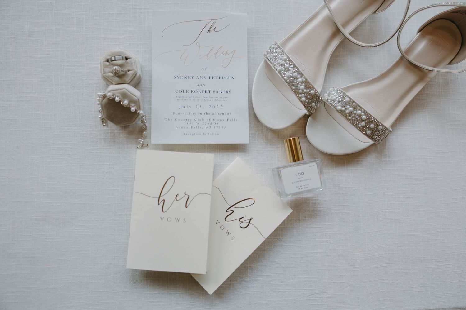 Elegant wedding details flatlay with invitations, rings, perfume and embellished bridal shoes.