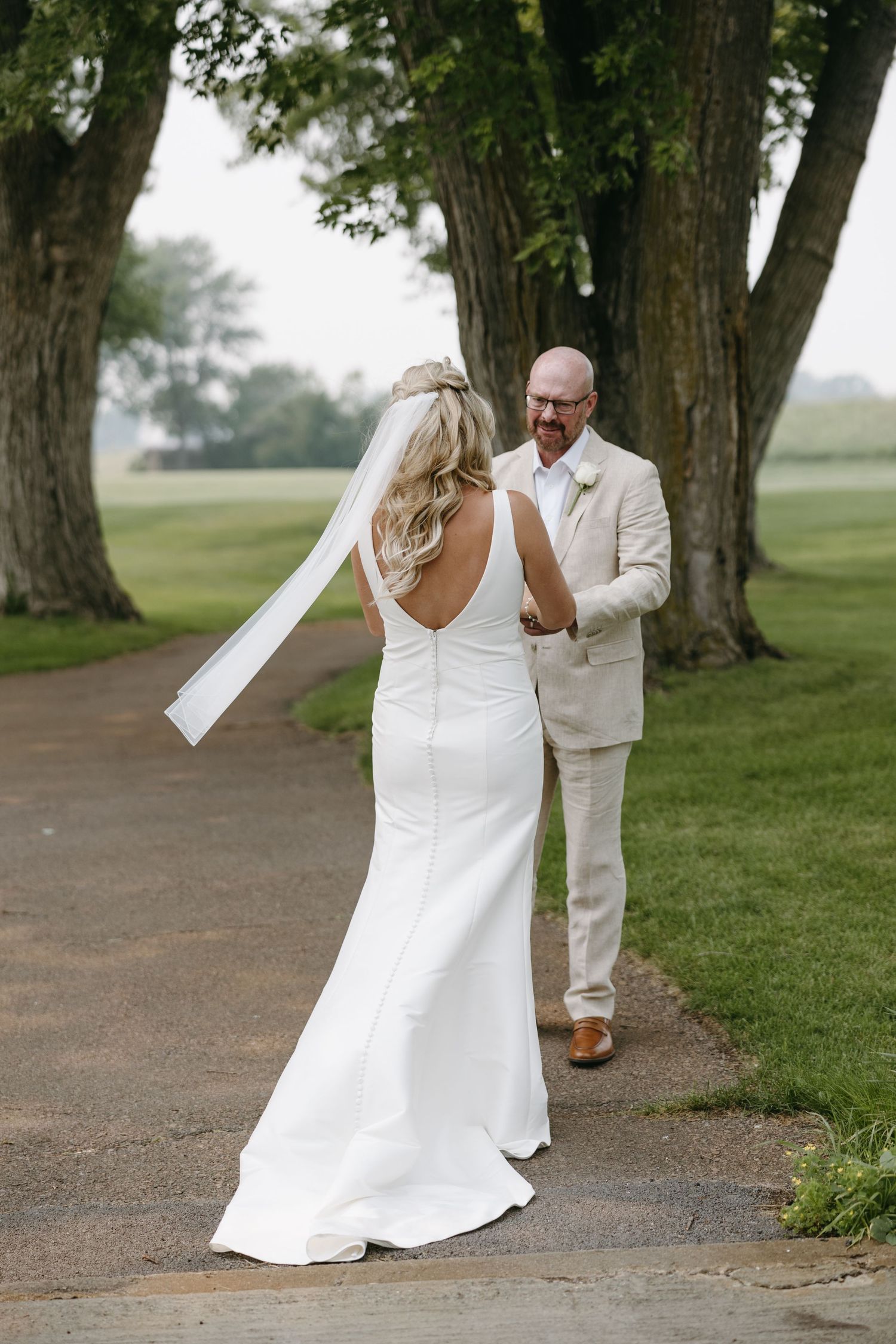 Bride in backless wedding dress walks path while groom in light suit looks on.