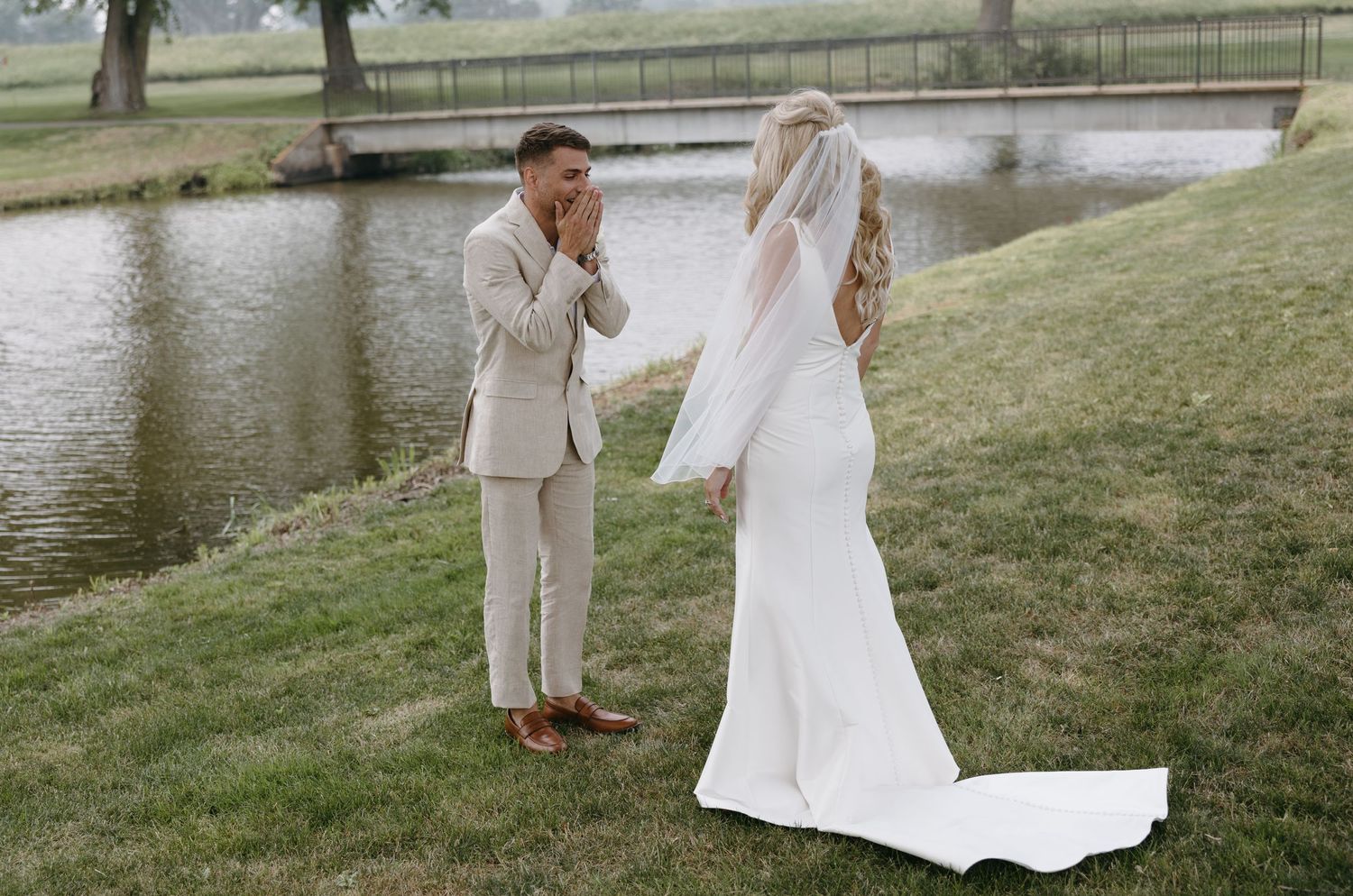 Emotional first look reaction between couple in light colored wedding attire by pond.