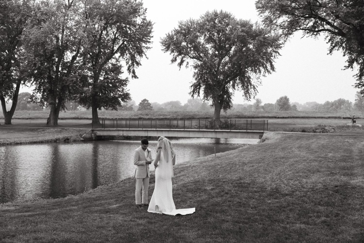 A serene black and white wedding photo by a pond with trees creating a peaceful backdrop.