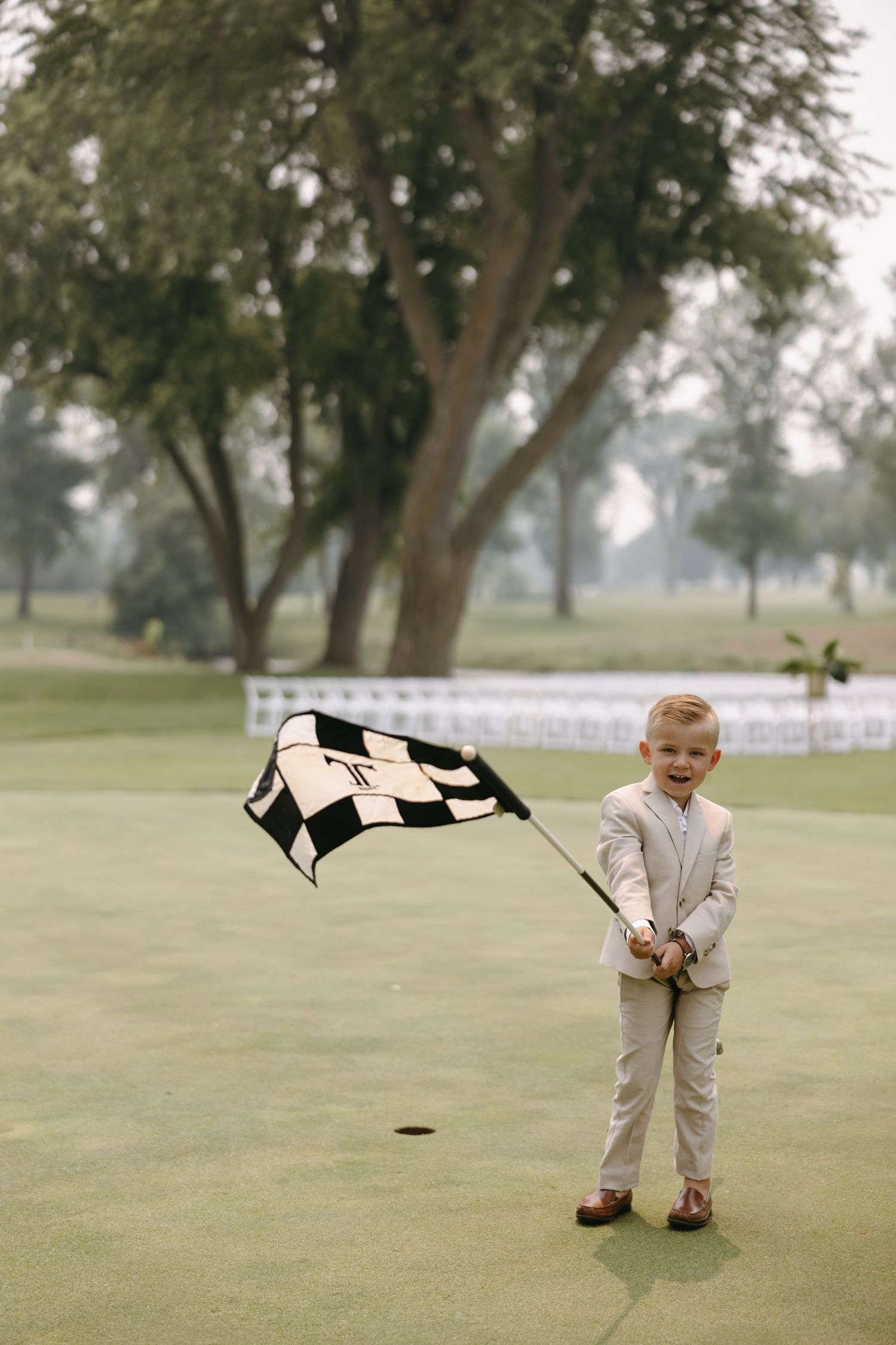 Young ring bearer in beige suit waves checkered flag on a golf course green during wedding ceremony.