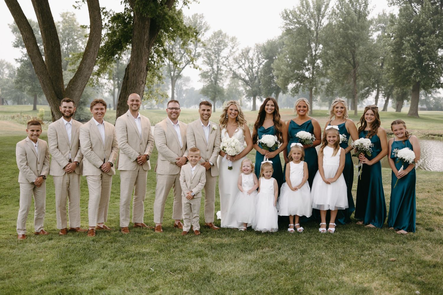 Wedding party poses on golf course with bridesmaids in teal dresses and groomsmen in beige suits.