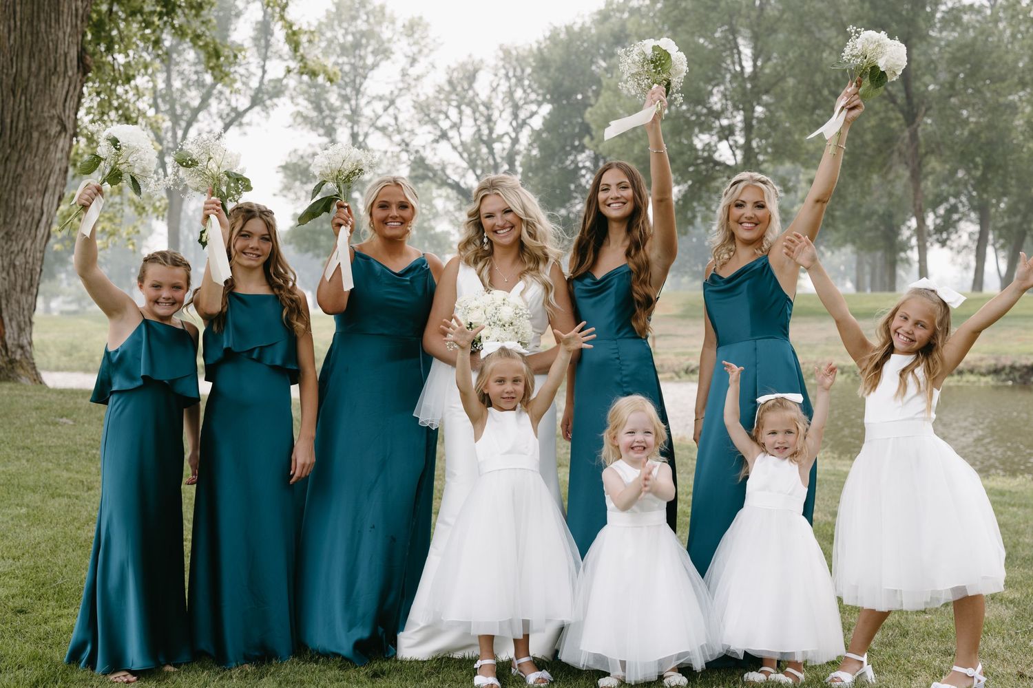 Bridal party in teal gowns and white dresses pose together outdoors for wedding photo.