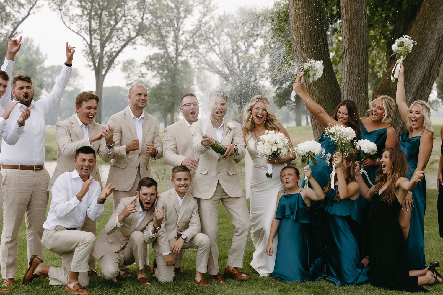 Joyful wedding party celebrates outdoors with bridesmaids in teal and groomsmen in beige.