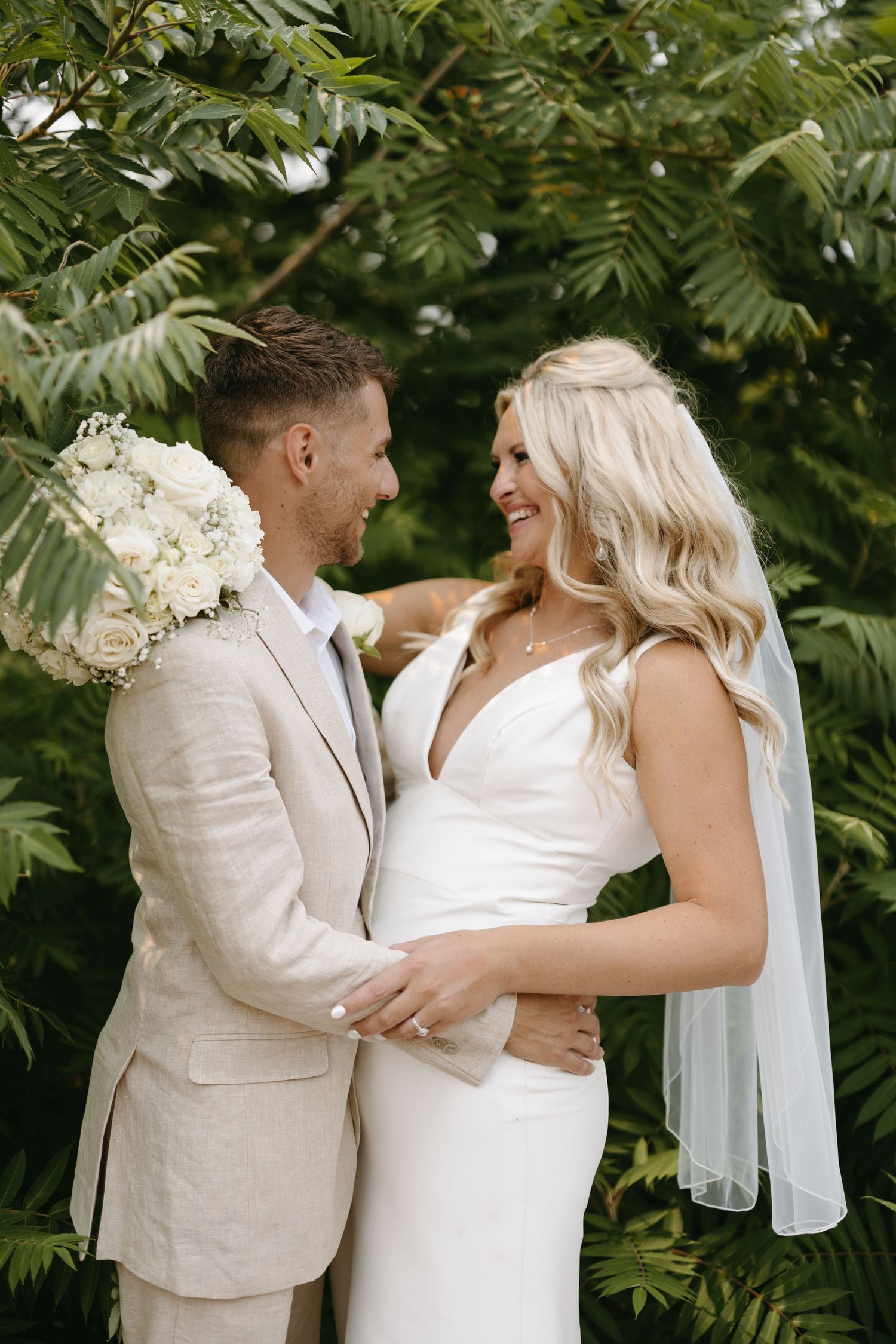 Newlyweds share loving glance against green foliage backdrop in elegant wedding attire.
