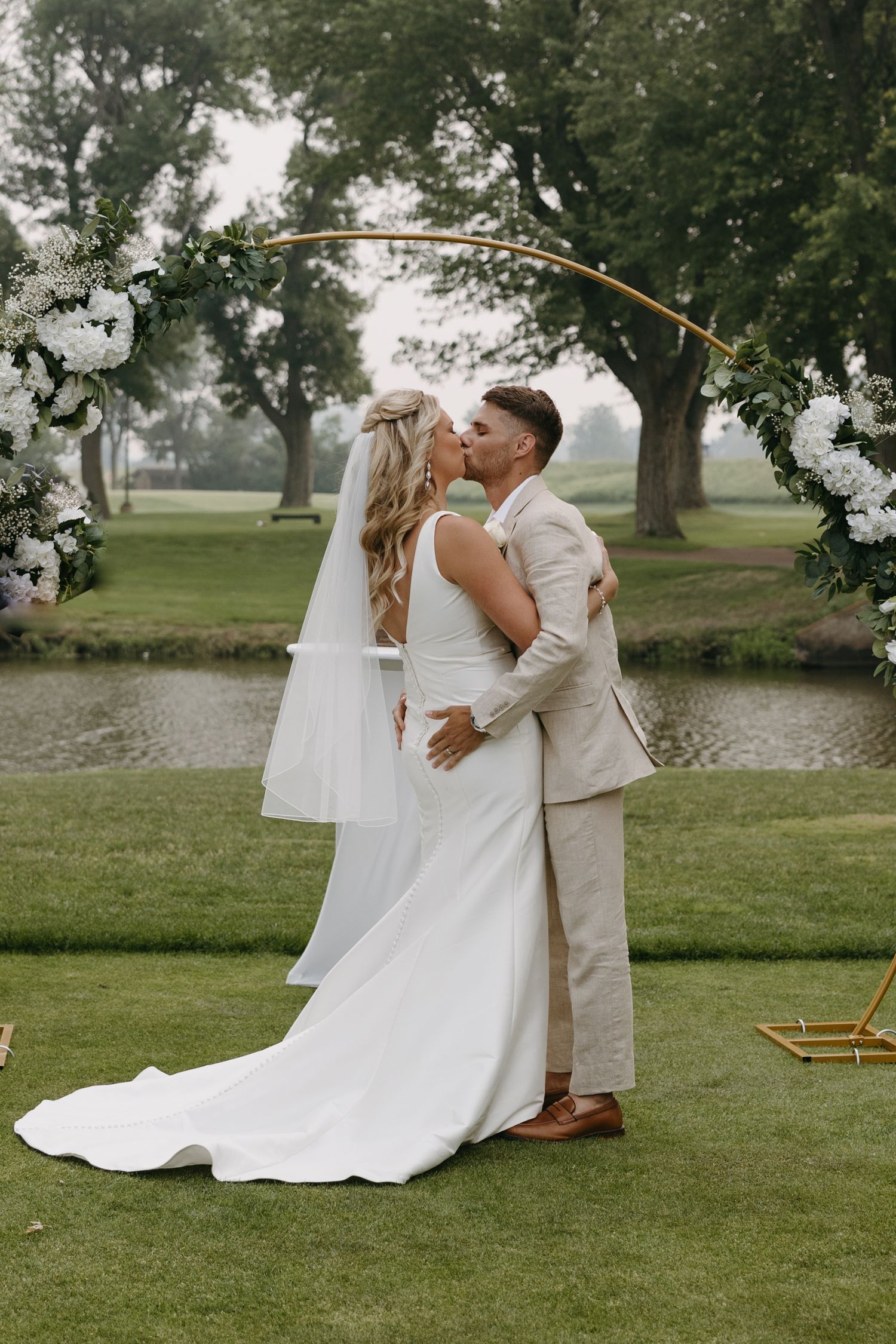 A romantic wedding moment under a floral arch on a misty golf course with a couple sharing their first kiss.