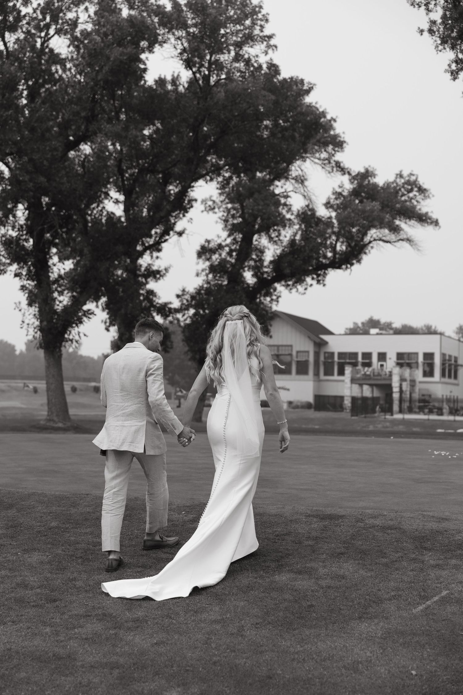 A black and white portrait of newlyweds walking hand in hand across a golf course.