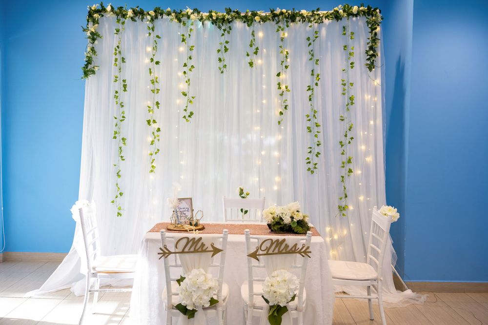 Wedding ceremony setup with white chairs and floral vine backdrop against a blue wall.