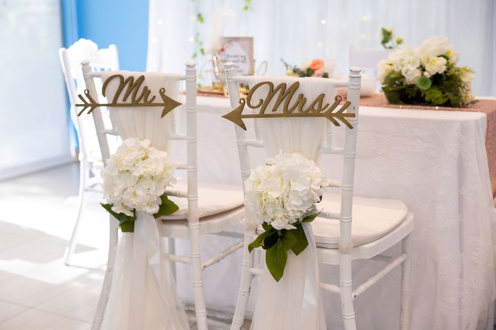 Elegant white wedding chairs decorated with 'Mr and Mrs' signs and floral arrangements in a sunlit venue.