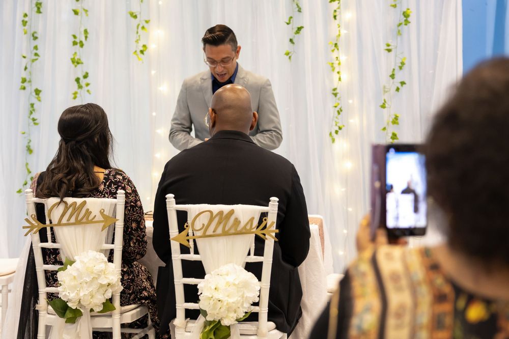 Intimate wedding ceremony setup with white floral backdrop and decorated chairs.