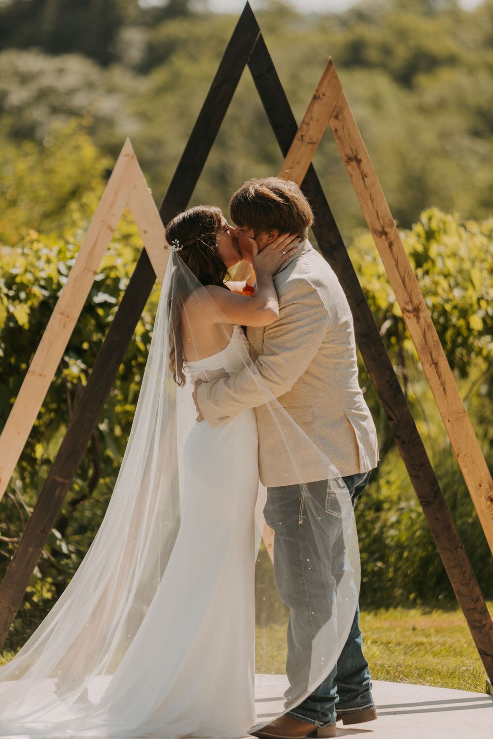 A romantic wedding couple shares an intimate kiss in a sunlit vineyard setting with a flowing white veil.