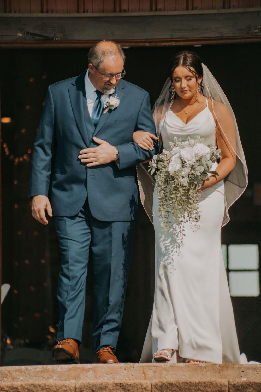 A bride in a white gown walks down steps arm-in-arm with someone in a blue suit during a wedding ceremony.