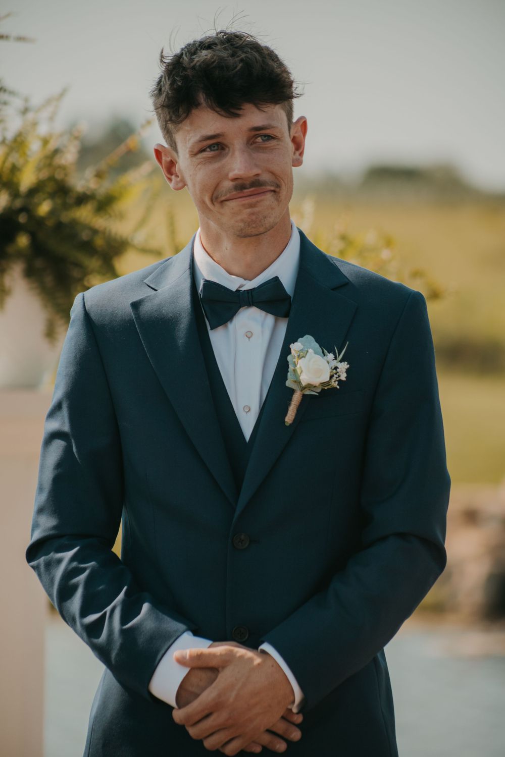 A person wearing a navy suit and bowtie with a floral boutonniere smiles before a wedding ceremony.