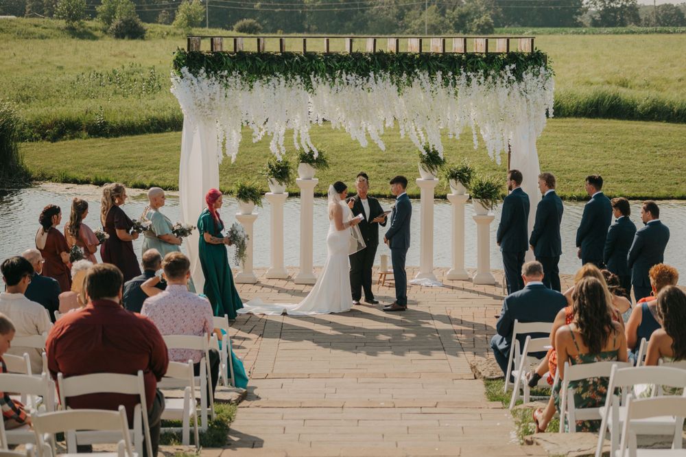 An outdoor wedding ceremony takes place under a white draped pergola with guests seated on white chairs.
