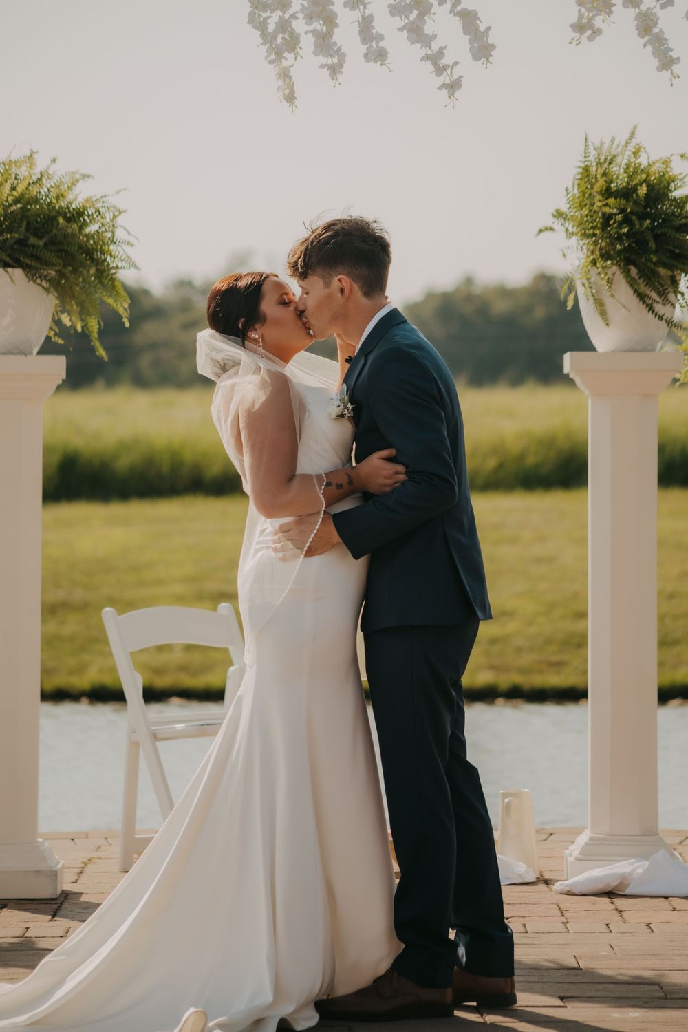 A couple shares an intimate moment between white columns with a lake view in the background.