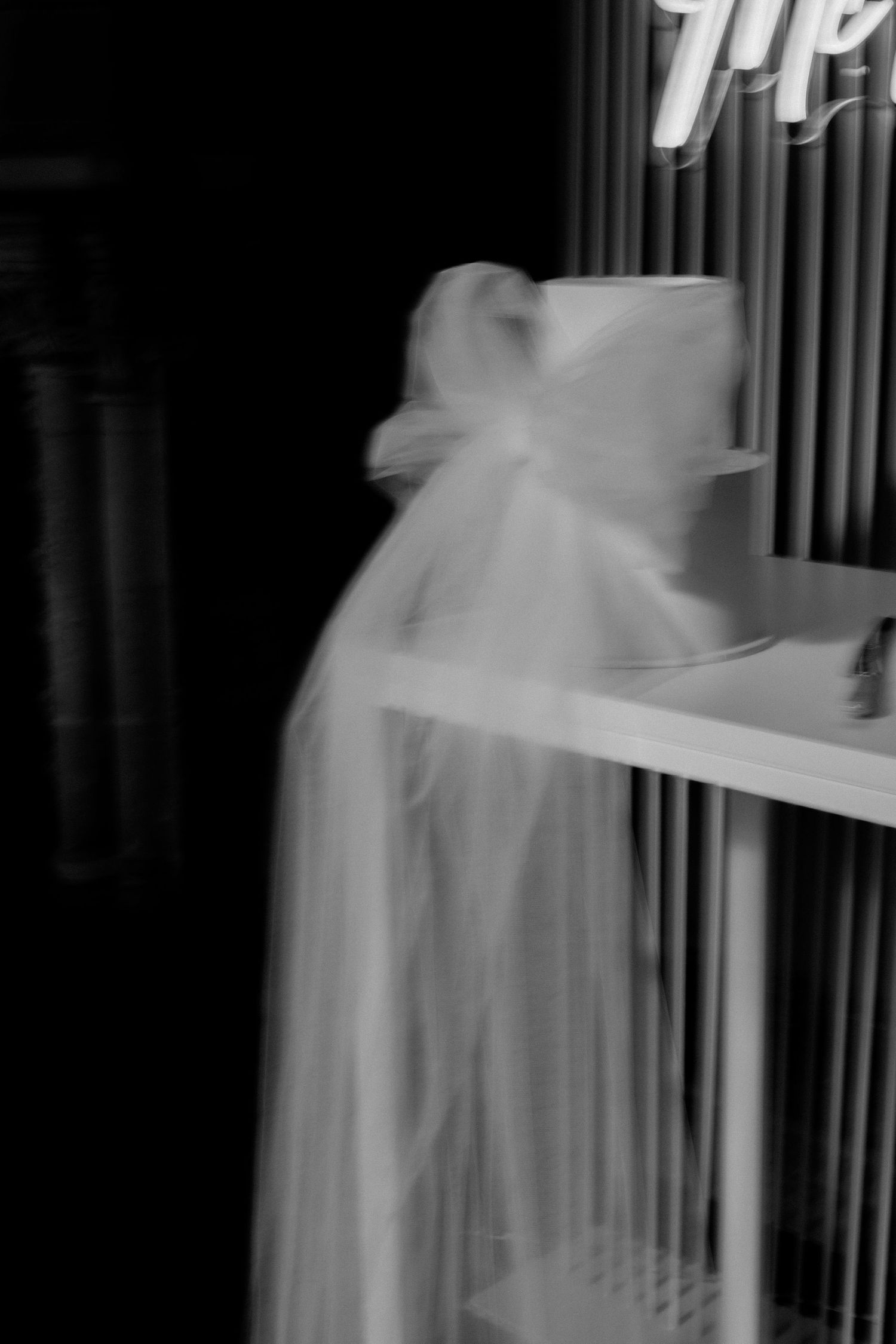 A blurry black and white photo of a ghostly figure in flowing white fabric standing near vertical bars or railings.