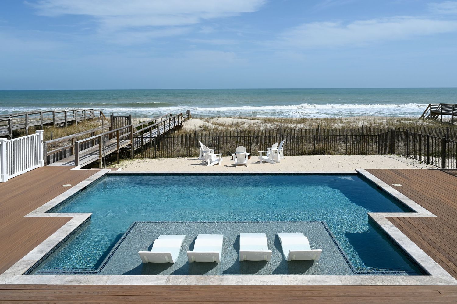 Modern beachfront pool with in-water loungers and ocean view beyond wooden boardwalk.