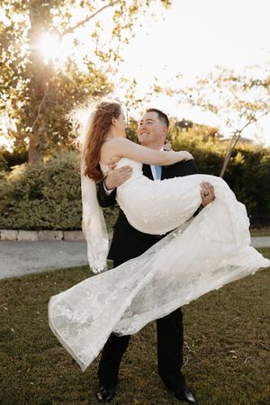 A romantic wedding couple embraces in golden sunlight as the groom carries the bride in her flowing lace dress outdoors.