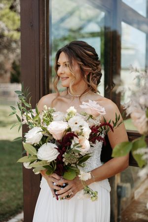 Elegant bridal bouquet featuring white roses, burgundy flowers, and greenery held against a flowing white wedding dress.