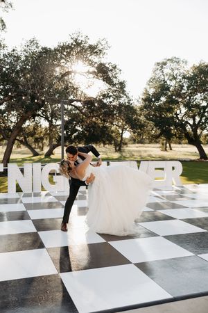 A couple dances on a giant outdoor chess board at sunset with trees in the background.
