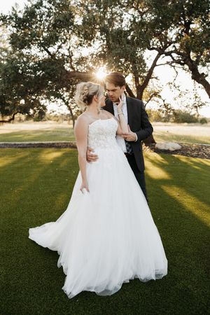 Romantic sunset wedding photo with couple dancing on lush green lawn as golden light streams through trees.
