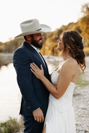 A romantic wedding couple embraces on a beach at sunset wearing a white dress and navy suit with cowboy hat.