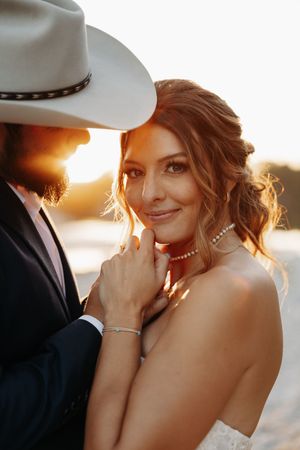 Bride in white dress and groom in cowboy hat share intimate moment at sunset.