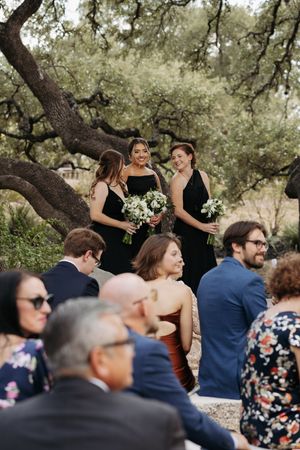Wedding guests seated outdoors under oak trees watching ceremony while bridesmaids in black dresses stand in background.
