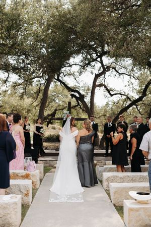Wedding ceremony guests gather outdoors under oak trees on a stone patio during a daytime celebration.