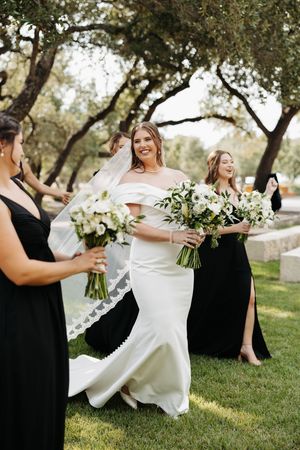 Bride and bridesmaids in black dresses hold white floral bouquets while posing together on grassy lawn at outdoor wedding.