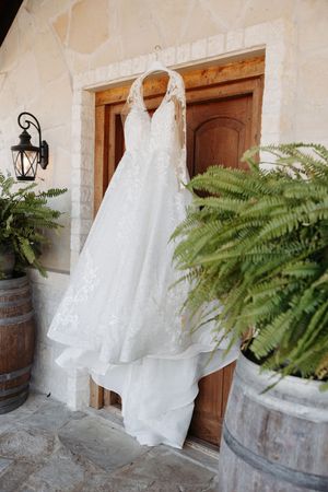 White wedding dress hanging on rustic stone wall with decorative fern plants and wooden barrels creating romantic ambiance.