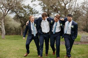 Groomsmen in navy suits walking and laughing together on a path surrounded by green grass and trees.