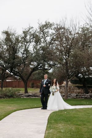 A wedding couple and officiant stand beneath sprawling oak trees on a stone pathway during an outdoor ceremony.