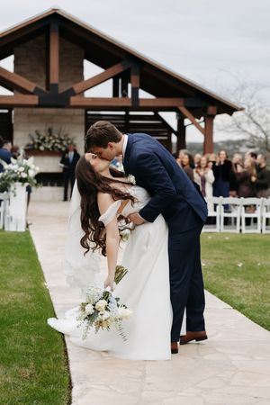 A romantic wedding couple shares a dip kiss on a path outside a rustic venue with guests watching in the background.