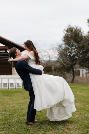 A romantic wedding couple embraces on a grassy lawn with white chairs and trees in the background.