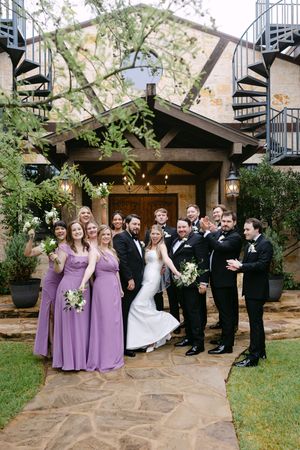 Wedding party poses outside rustic venue, bridesmaids in purple gowns, groomsmen in tuxedos, bride in white gown holding bouquet.