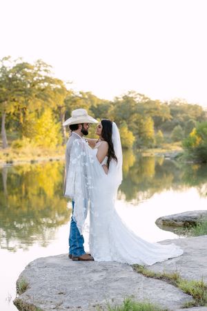 Bride and groom share a romantic moment by a reflective lake, she in a white gown, he in a cowboy hat and jeans.